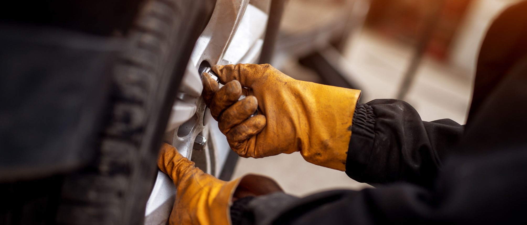 An experienced mechanic with orange gloves is putting screws on a placed wheel on a car.