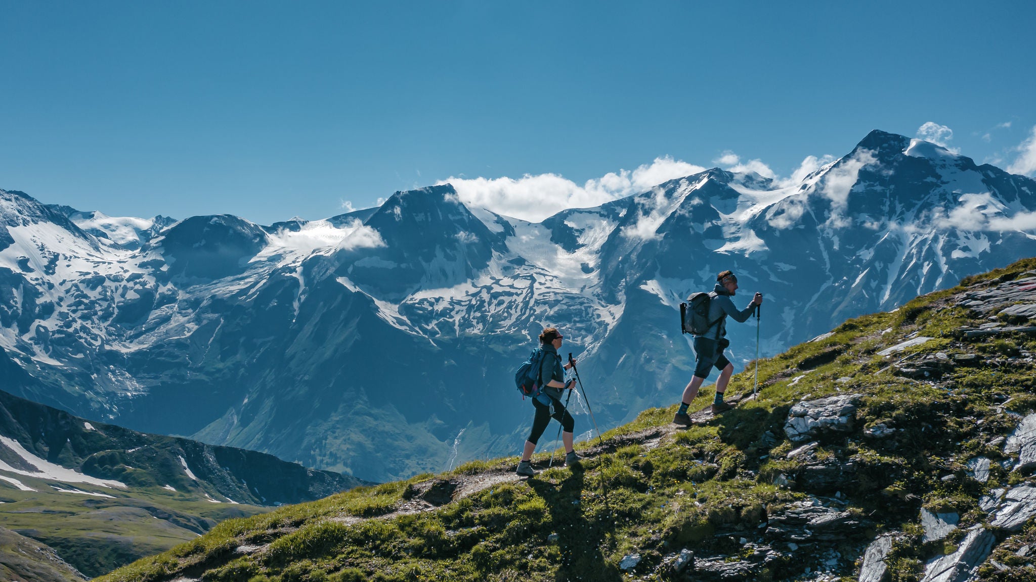 Two young hikers (a couple) walking up a mountain in Austria in summer, with scenic snow covered mountains on the background