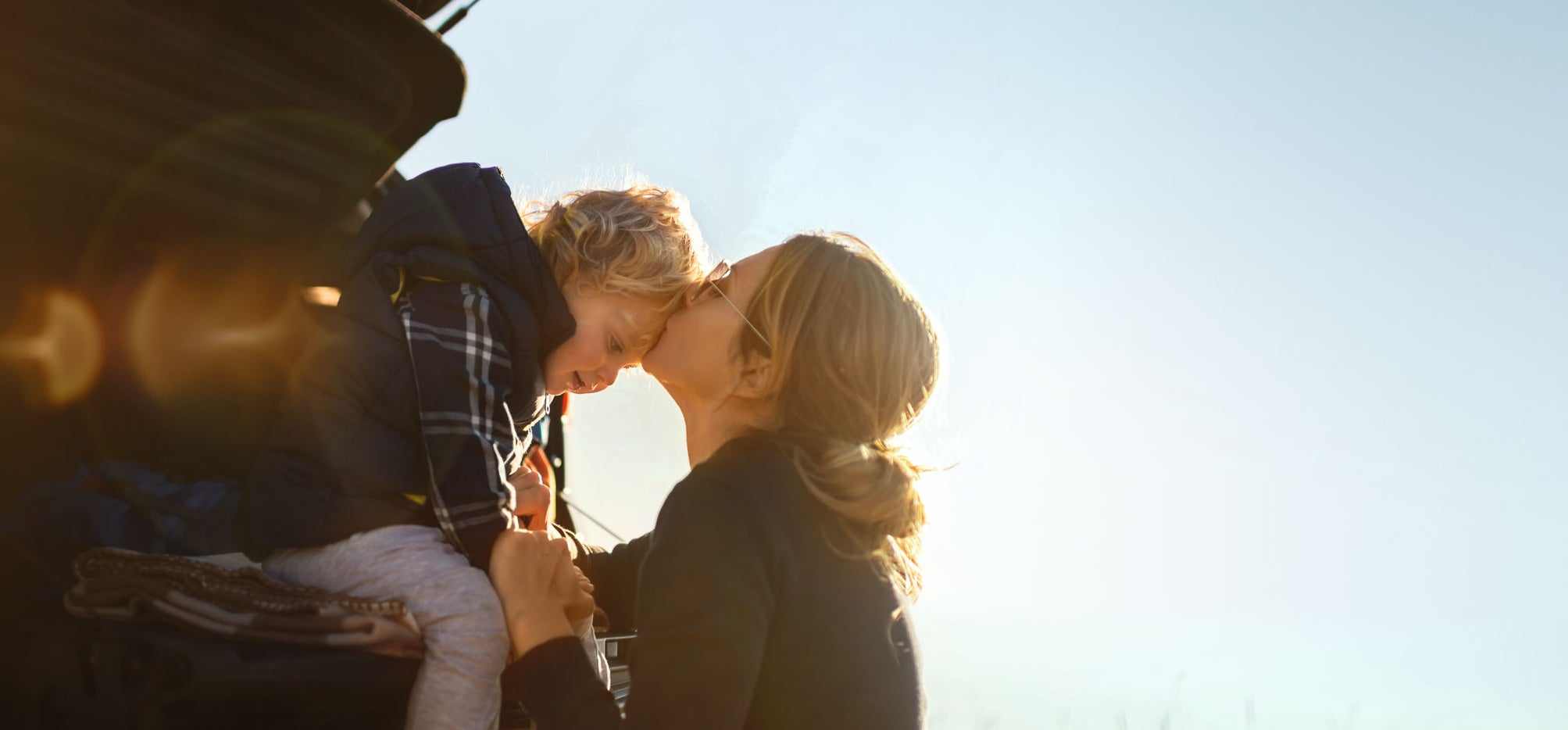 Mother and little son travel by car in mountains.Little boy sitting in car trunk while they makes a break and mother gives him a kiss.