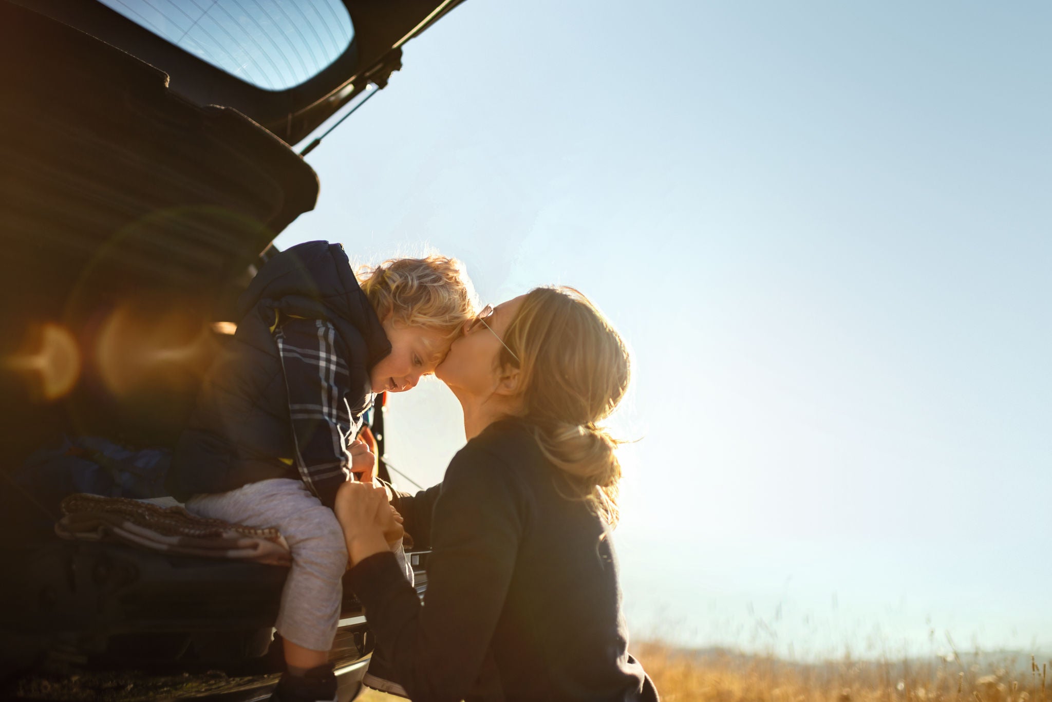 Mother and little son travel by car in mountains.Little boy sitting in car trunk while they makes a break and mother gives him a kiss.